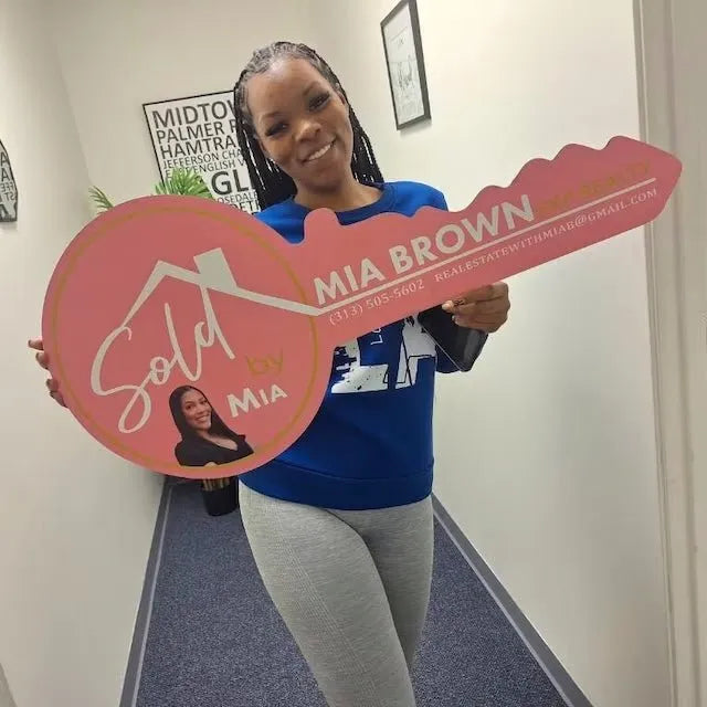 Woman holding large pink key sign reading Sold by Mia Brown with real estate contact details indoors