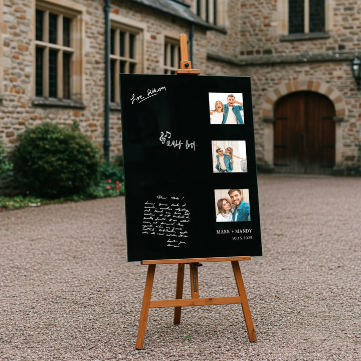 Black wedding guest book sign with photos and handwritten messages on wooden easel outside stone building