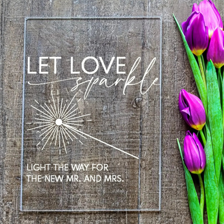 Clear wedding table sign with white text "Let Love Sparkle" and purple tulips on rustic wooden surface