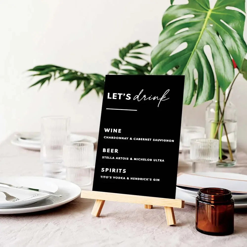 Black wedding bar menu sign with white text on wooden easel, surrounded by tableware and green leaves