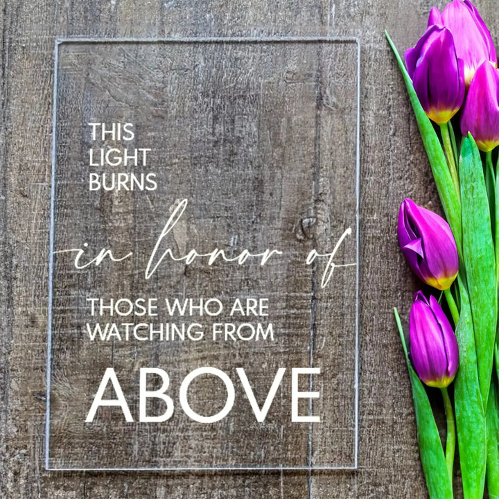 A clear acrylic memorial sign with white text and purple tulips on a wooden surface