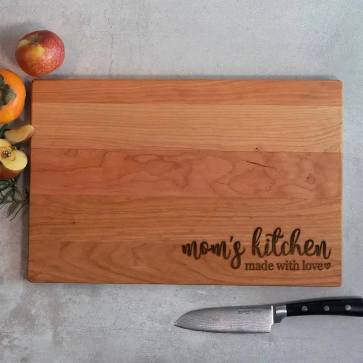 Personalized wood cutting board engraved with 'mom's kitchen made with love', knife and fruit nearby.