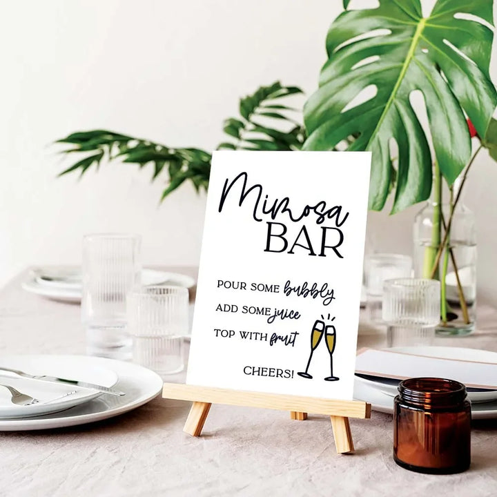 Mimosa Bar sign on wooden easel with champagne recipe on table set for event with green leaves