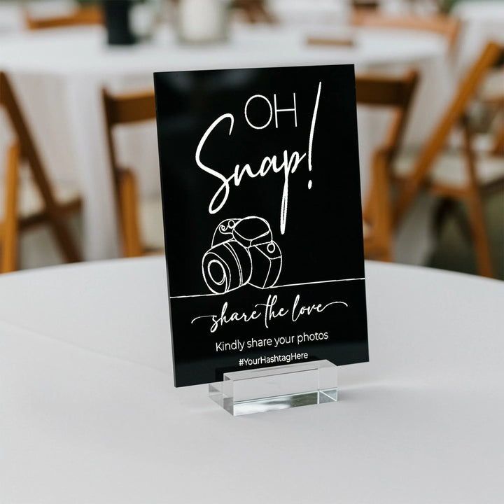Black wedding table sign with white camera graphic and text encouraging photo sharing with hashtag