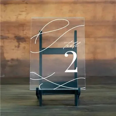 Ornate acrylic table number 2 sign with elegant white script on black easel against wooden background