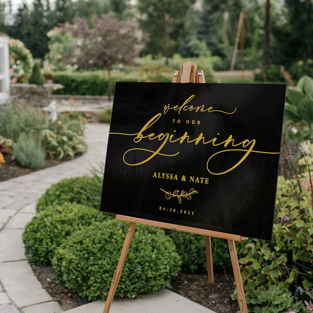 Black wedding welcome sign with gold script reading 'Welcome to our beginning Alyssa & Nate 04.28.2023' on wooden easel in garden path