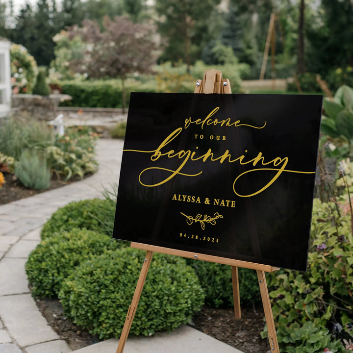 Black wedding welcome sign with gold script reading 'Welcome to our beginning Alyssa & Nate 04.28.2023' on wooden easel in garden path