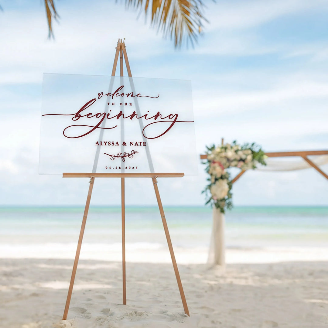 Acrylic wedding welcome sign with cursive text on wooden easel on sandy beach with floral wedding arch background