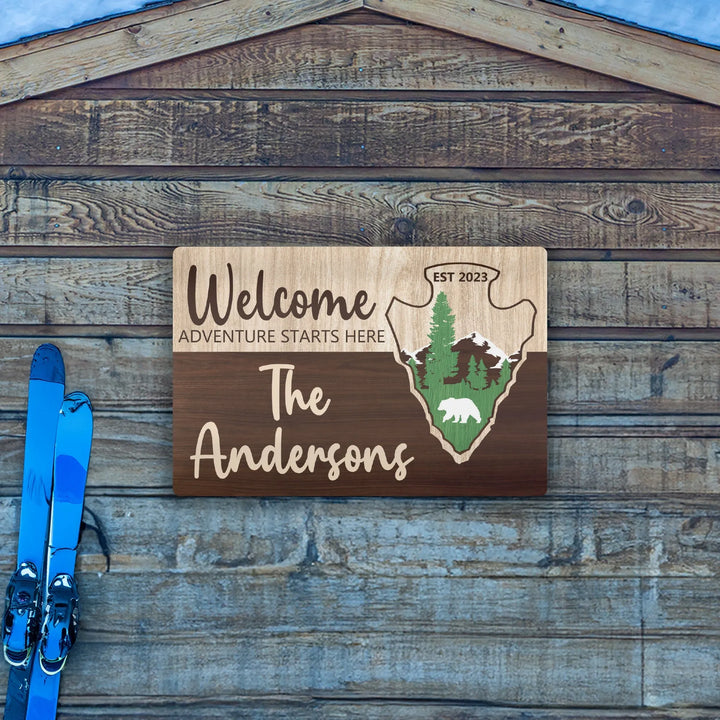 Personalized wooden welcome sign with adventure theme, mountains, trees, bear, and blue skis on wooden wall