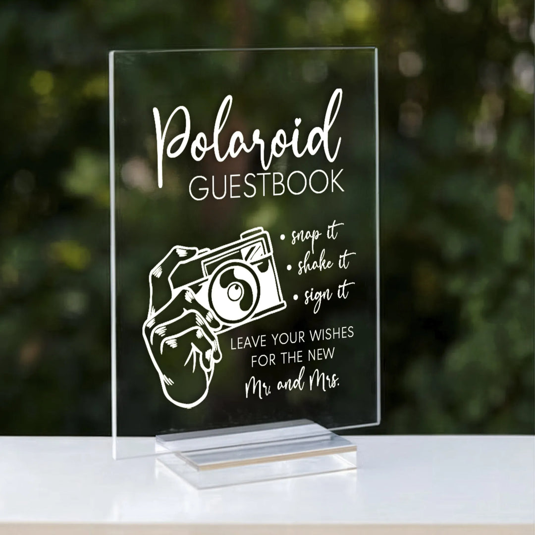 Clear acrylic Polaroid guestbook sign with white text and hand holding camera illustration on stand