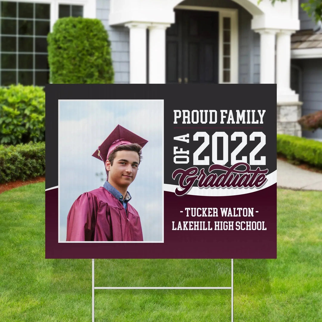 Yard sign on lawn with photo of male 2022 graduate in maroon cap and gown, text Proud Family of a 2022 Graduate