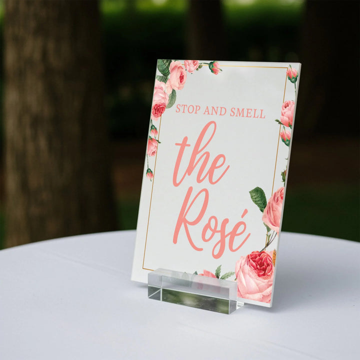Elegant wedding table sign with pink roses and text 'Stop and smell the Rosé' on white tablecloth