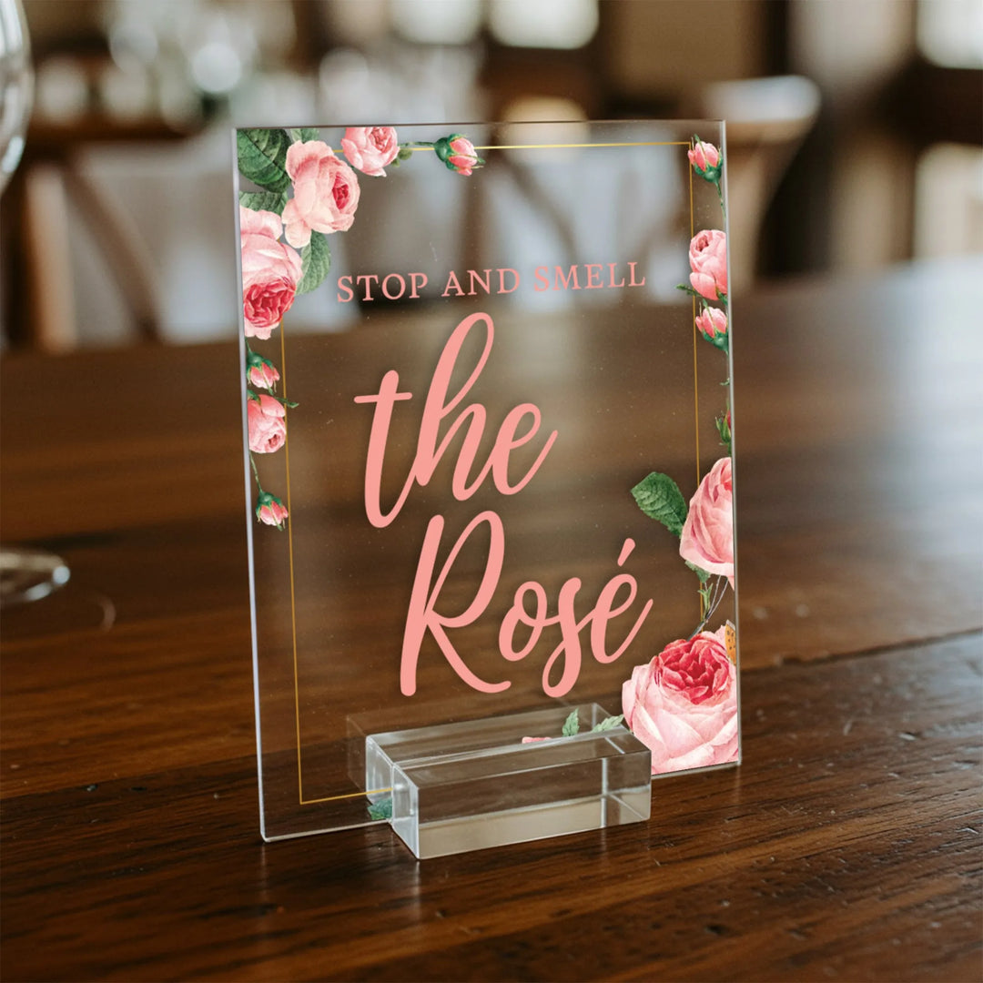 Acrylic wedding table sign with pink rose floral design and text 'Stop and smell the Rosé' on wooden surface