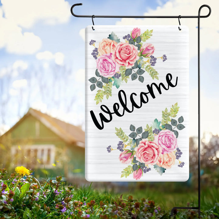 Metal garden flag with watercolor pink roses and green leaves, black welcome text, outdoor yard setting