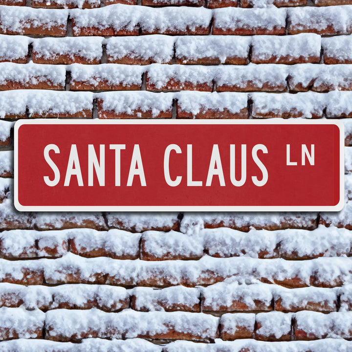 Red metal street sign reading Santa Claus Ln on snow-covered brick wall