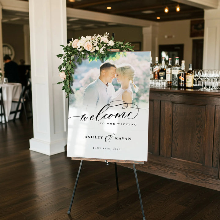 Elegant wedding welcome sign with couple photo, floral arrangement, and bar in background