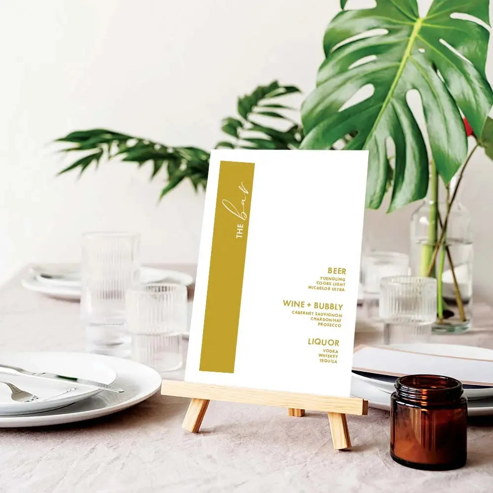 Elegant wedding bar menu sign with gold stripe on wooden easel, glassware and green tropical leaves