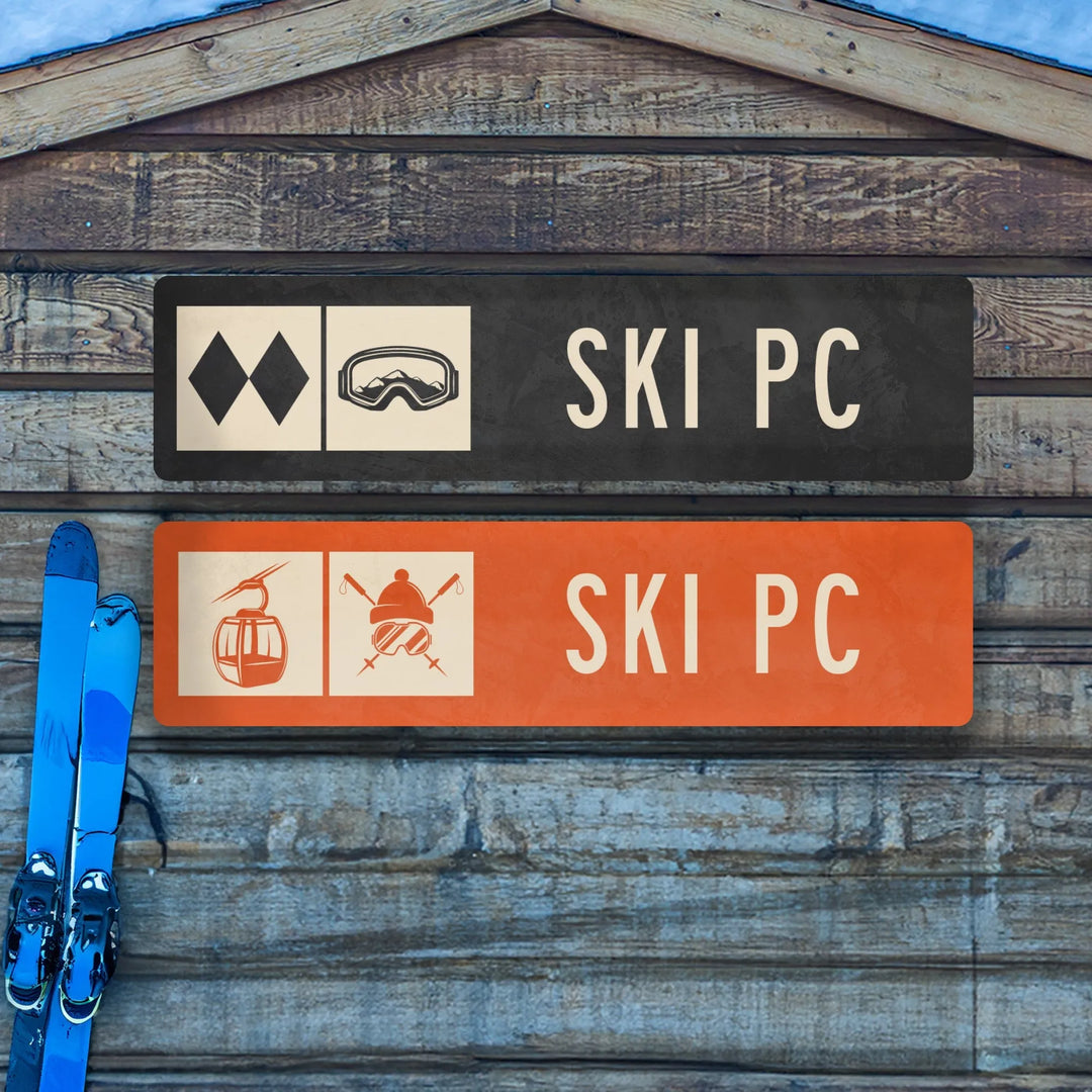 Two metal ski trail signs on wooden wall with blue skis beside them, one black with goggles, one orange with cable car