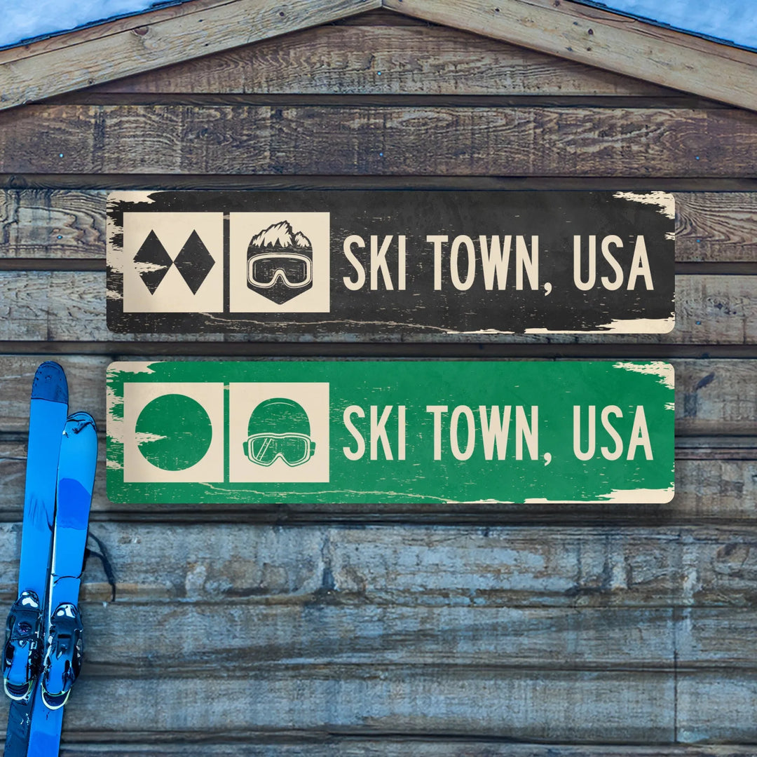 Two distressed metal signs on wood wall, black and green, reading Ski Town USA with ski symbol and goggles icon, blue skis nearby