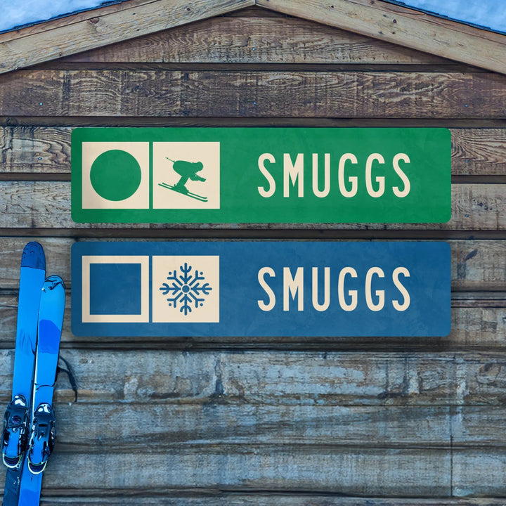 Two metal ski trail signs mounted on wood: green with skier icon, blue with snowflake, both labeled Smuggs