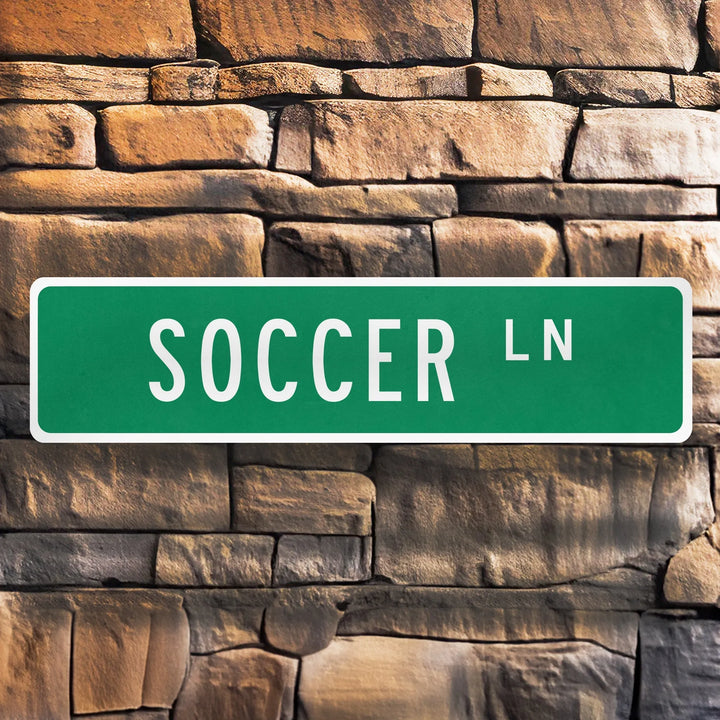 Green metal street sign with white text reading Soccer Ln mounted on a textured stone wall