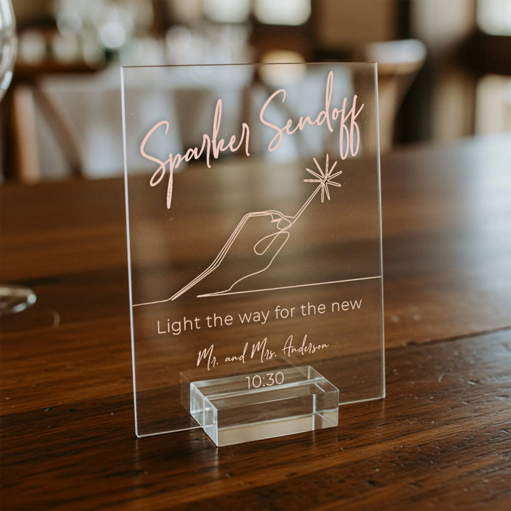 Clear acrylic wedding table sign reading Sparkler Sendoff with hand holding sparkler illustration on wooden surface