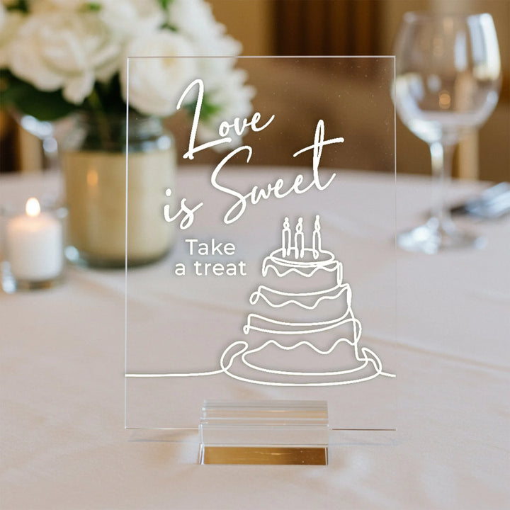 Clear acrylic wedding sign with white cake illustration and text Love is Sweet, Take a treat on a table
