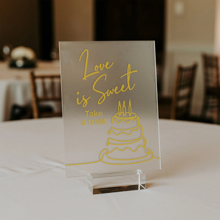 Clear acrylic wedding sign with yellow text "Love is Sweet, Take a treat" and cake illustration on white table