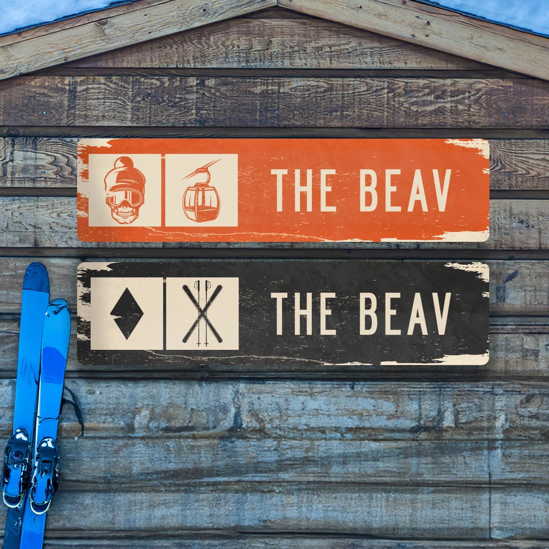 Distressed metal ski trail signs labeled The Beav with orange and black backgrounds, next to blue skis on wooden wall