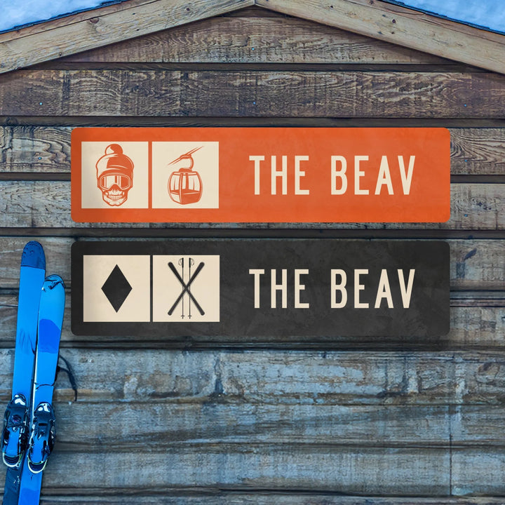 Two metal ski trail signs labeled The Beav with ski and gondola icons on wooden wall and blue skis nearby