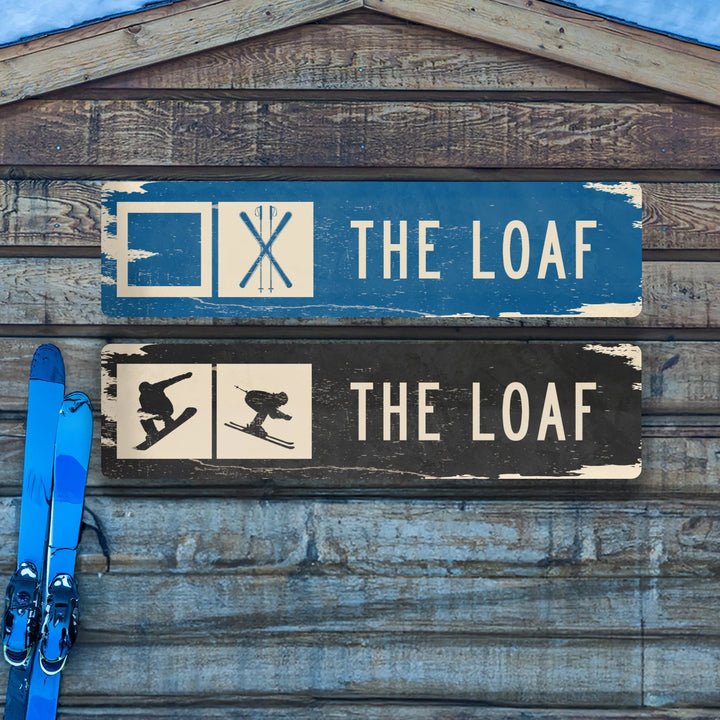 Distressed metal ski trail signs labeled The Loaf with ski and snowboard icons on wooden wall with blue skis