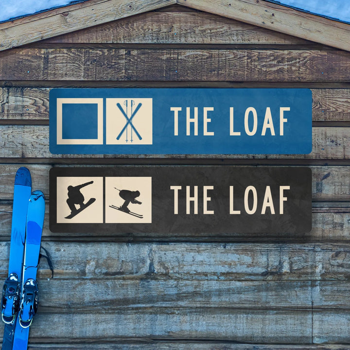 Two metal ski trail signs reading The Loaf with ski poles and silhouettes of snowboarder and skier on wooden background