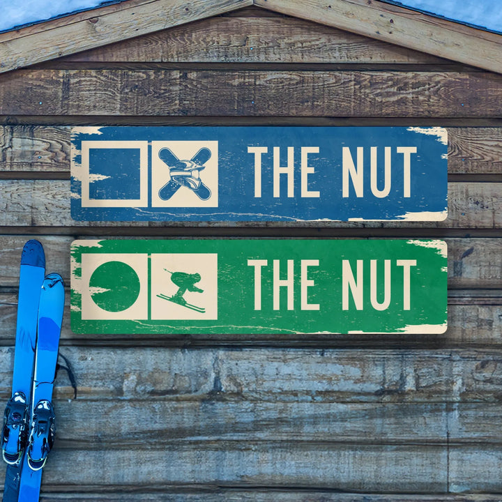 Two distressed metal ski trail signs labeled The Nut with skier and snowboarder icons on wooden wall, blue skis nearby