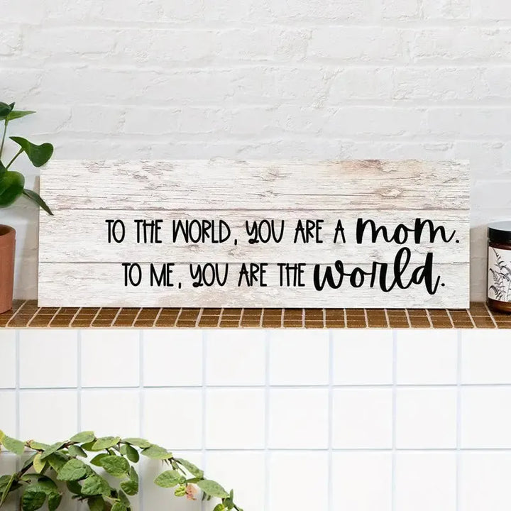 Rustic wood sign with 'To the world, you are a mom...' quote on tiled shelf with plants.