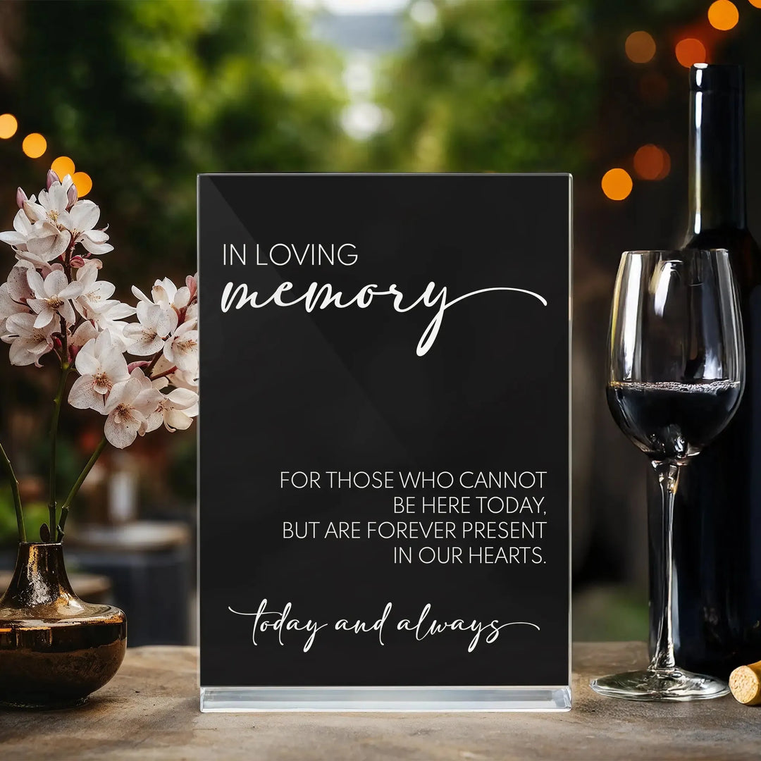 Black memorial wedding table sign with white text, pink flowers in vase, and wine glass with bottle