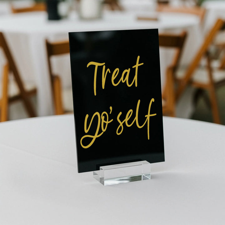 Black wedding table sign with gold script text 'Treat yo'self' on white table with wooden chairs