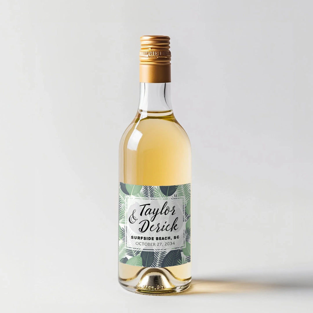 Mini wine bottle with gold cap and tropical leaves wedding label on white background
