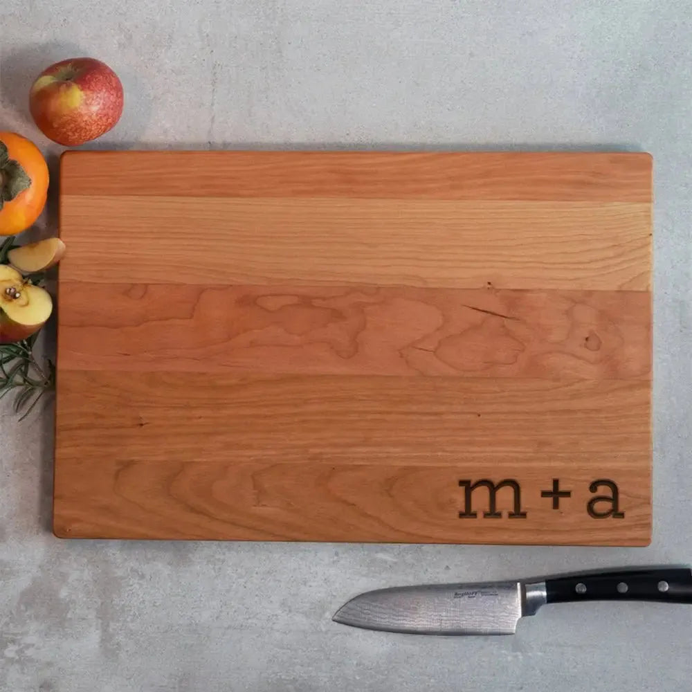 Personalized wood cutting board with 'm + a' engraving, chef knife, apples and persimmon on concrete.