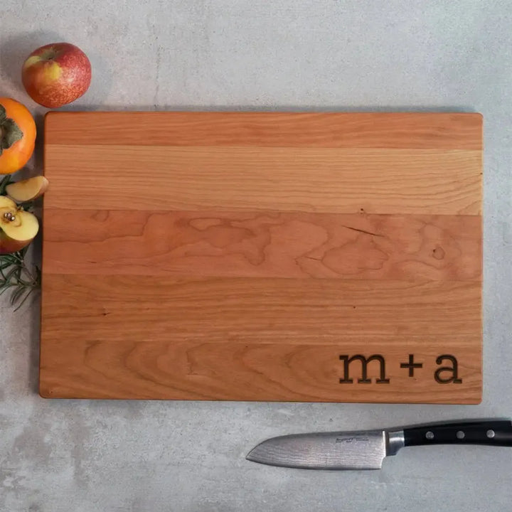 Personalized wood cutting board with 'm + a' engraving, chef knife, apples and persimmon on concrete.