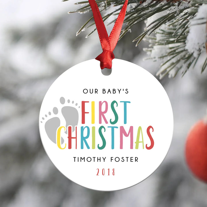 Colorful baby first Christmas ornament with footprints hanging on snowy pine tree