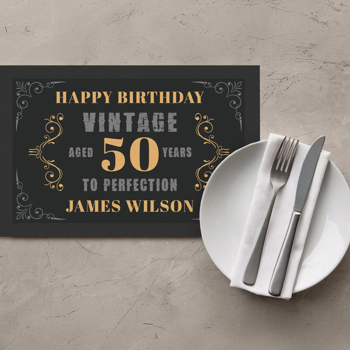 Vintage 50th birthday placemat with ornate gold accents beside a plate and cutlery