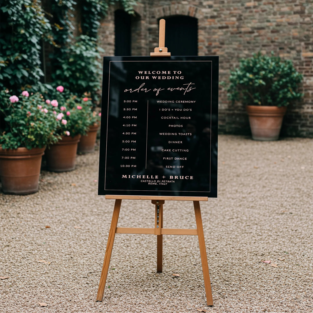 Elegant black wedding day-of-events timeline sign on wooden easel in outdoor garden venue with plant pots