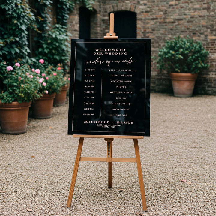 Elegant black wedding day-of-events timeline sign on wooden easel in outdoor garden venue with plant pots