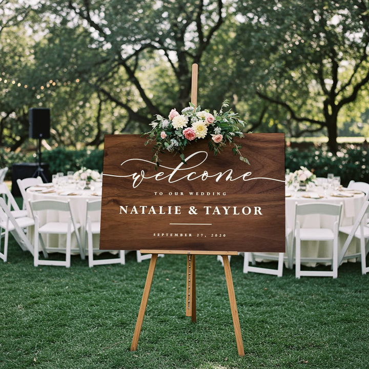 Rustic wooden wedding welcome sign with floral arrangement, outdoor reception with white chairs and tables